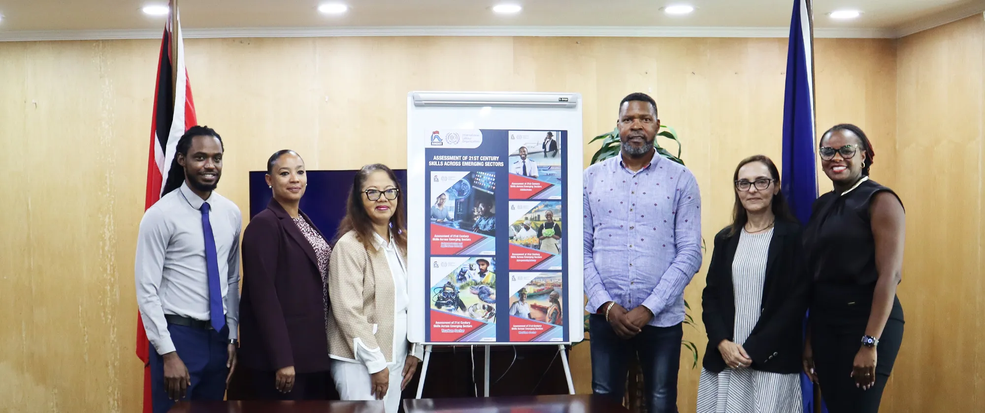 Seven persons standing in a conference room at the ILO Office in Port of Spain, Trinidad and Tobago, beside a display board showcasing the Assessment of 21st Century Skills Across Emerging Sectors research reports.