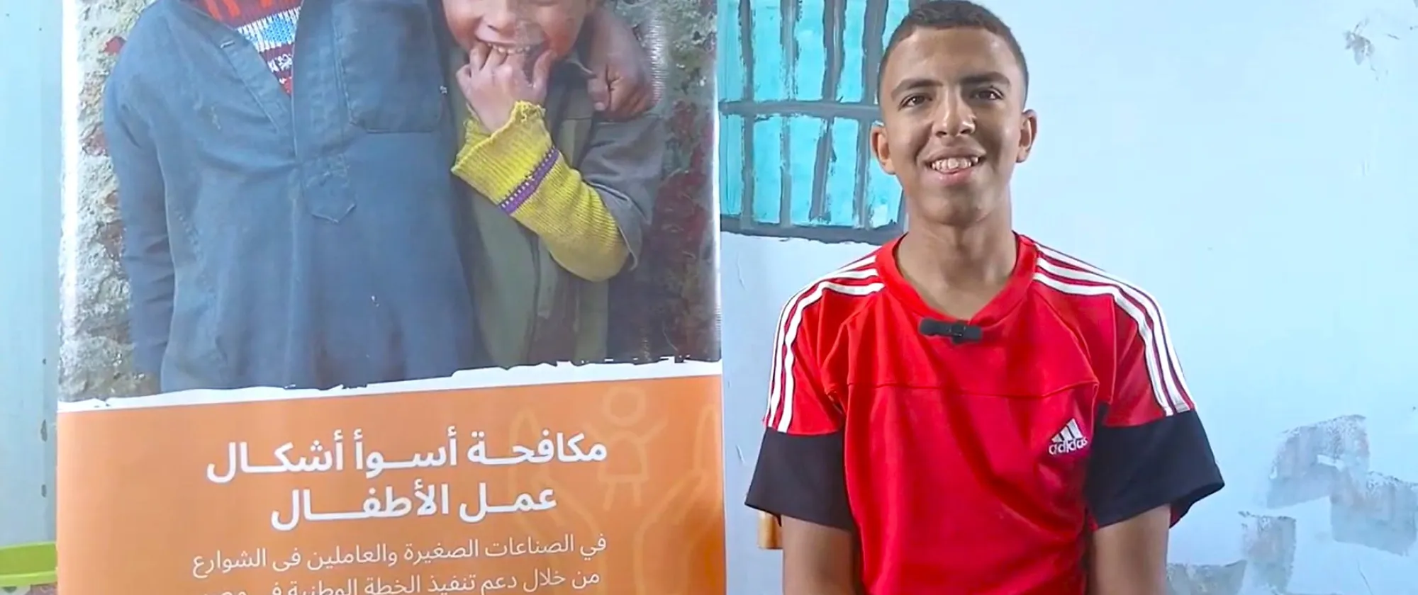 Young boy smiles while standing next to a poster on combatting the worst forms of child labour.