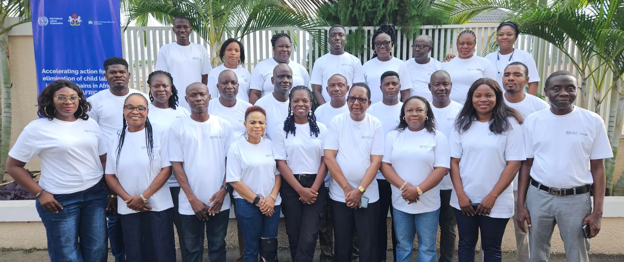 A group of newly certified cooperative trainers standing together outdoors in matching white shirts with an ILO banner displayed beside them