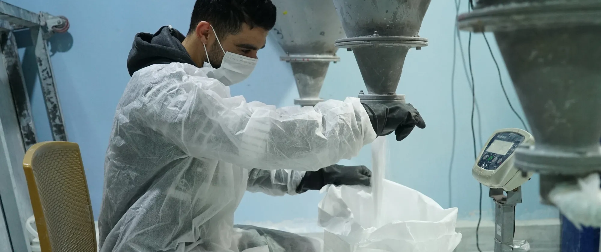 Worker filling a bag with powder from an industrial mixing machine while wearing protective equipment. Sahab, Jordan - June 2022