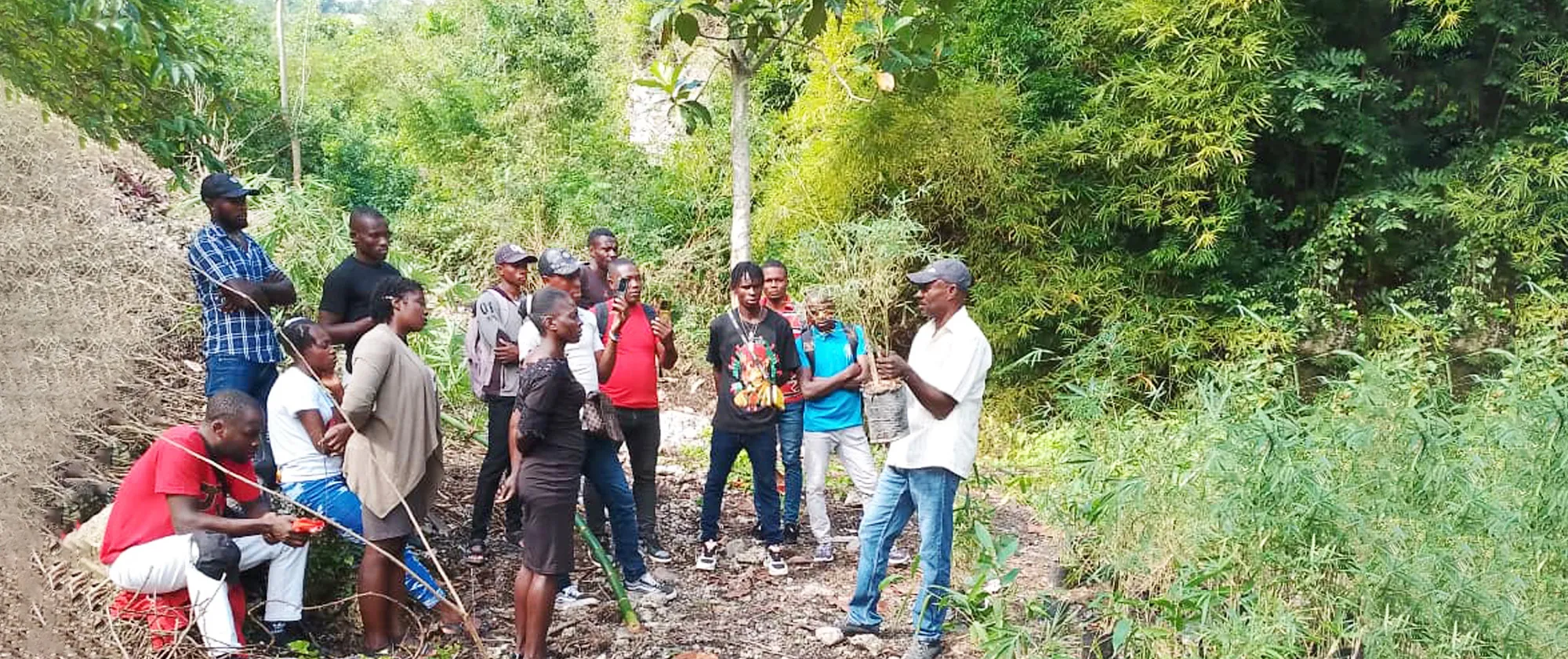 A group of approximately 15-20 people gather outdoors in a rural, vegetated area in Jeremie Haiti for a green skills training session. An instructor in light-coloured clothing and a cap stands on the right, gesturing while teaching the assembled participants. 