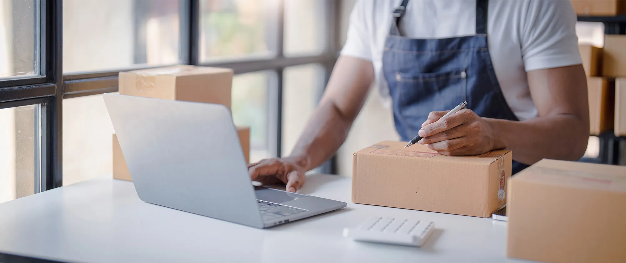 A person typing on a laptop while writing on a cardboard shipping box, with several packaged boxes around them.