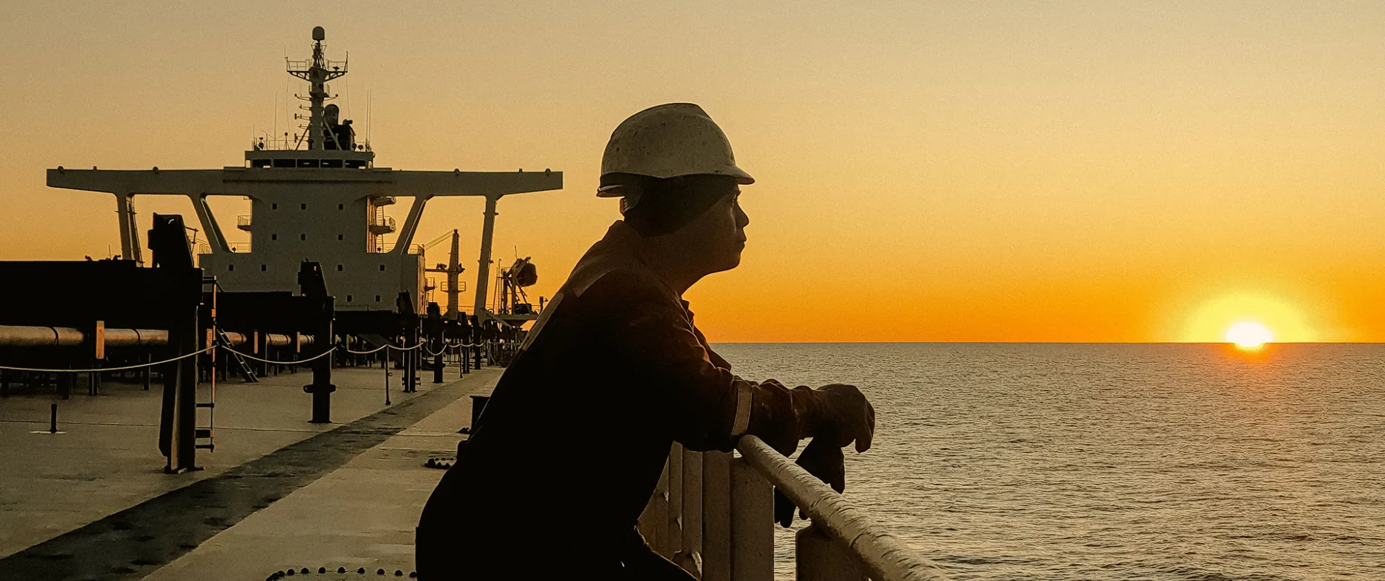 Seafarer standing at the railing of a cargo vessel, gazing at the horizon as the sun sets over the ocean