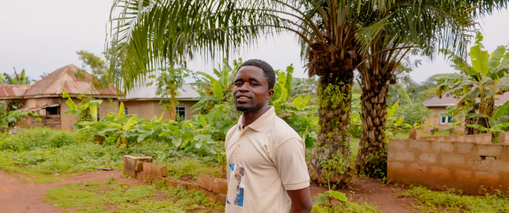 A young Nigerian man, Orifa Taiwo, stands outdoors in his village in Ondo State, surrounded by greenery and family homes, looking calmly towards the camera.