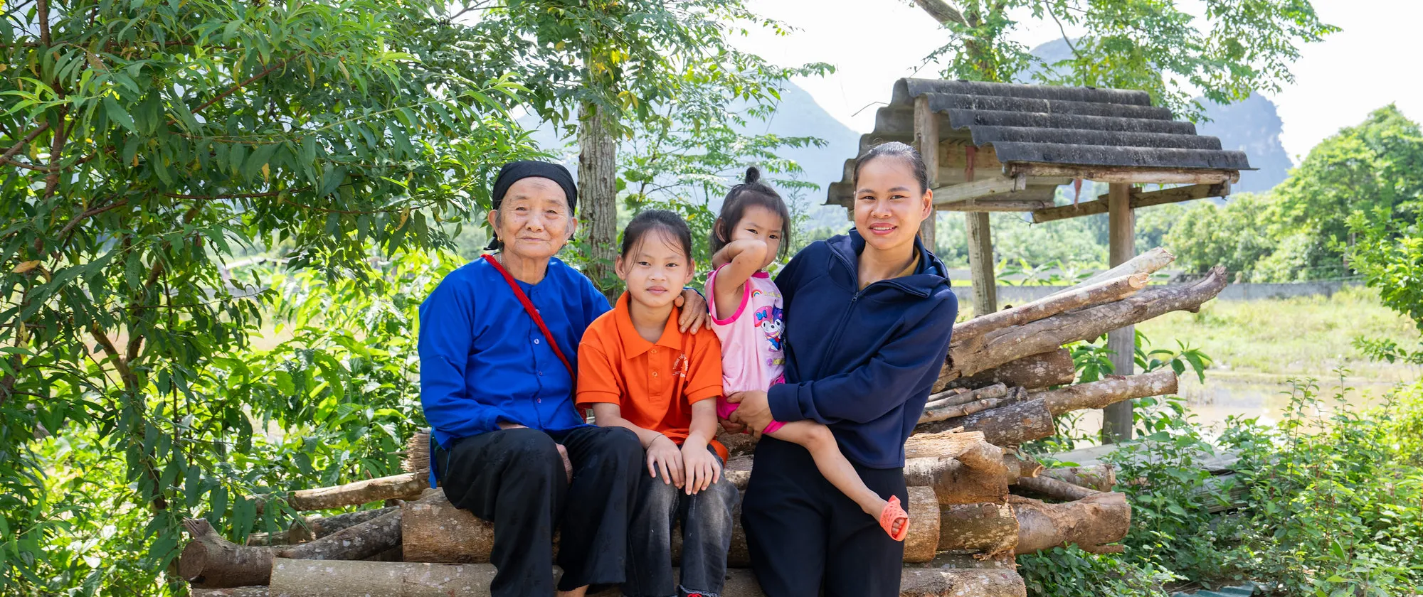 A woman holding a young child stands beside an older child and an elderly woman, all seated in front of stacked logs.