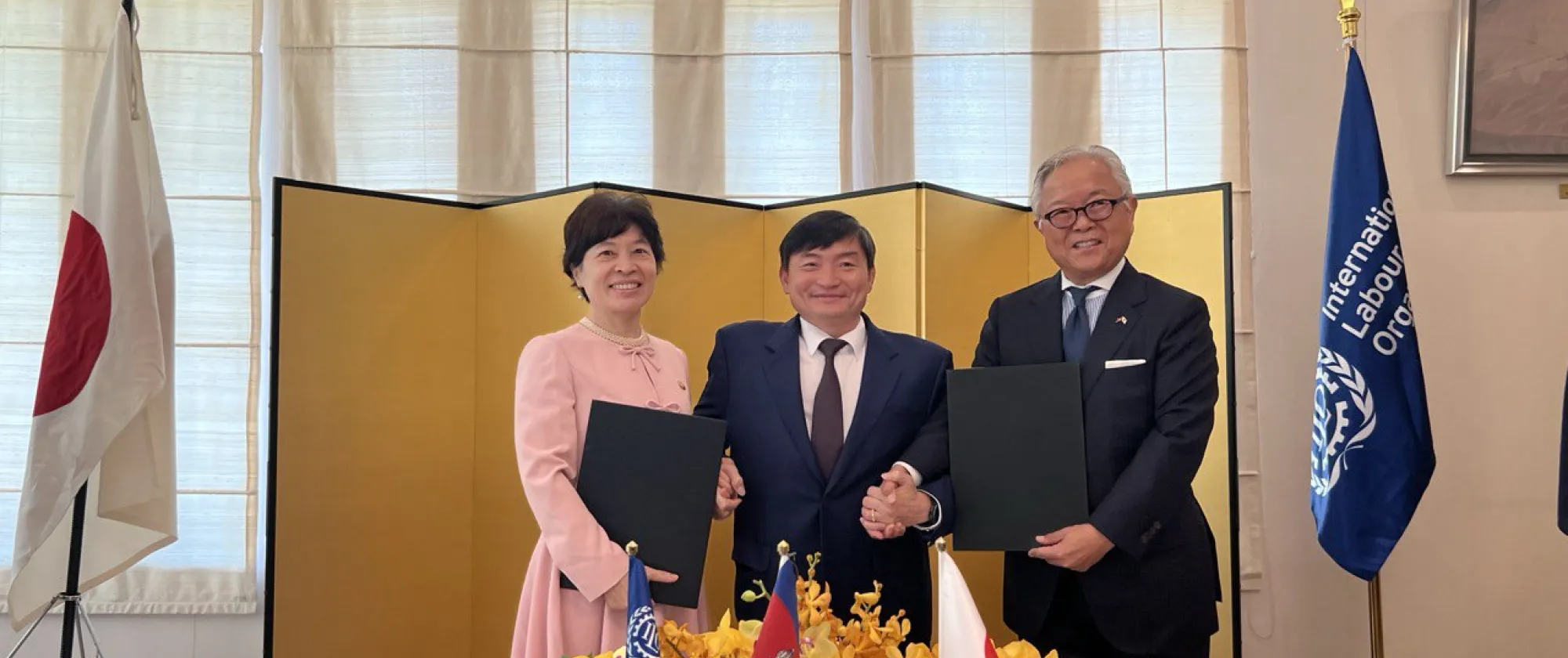 Three people stand between the flags of Japan and ILO.