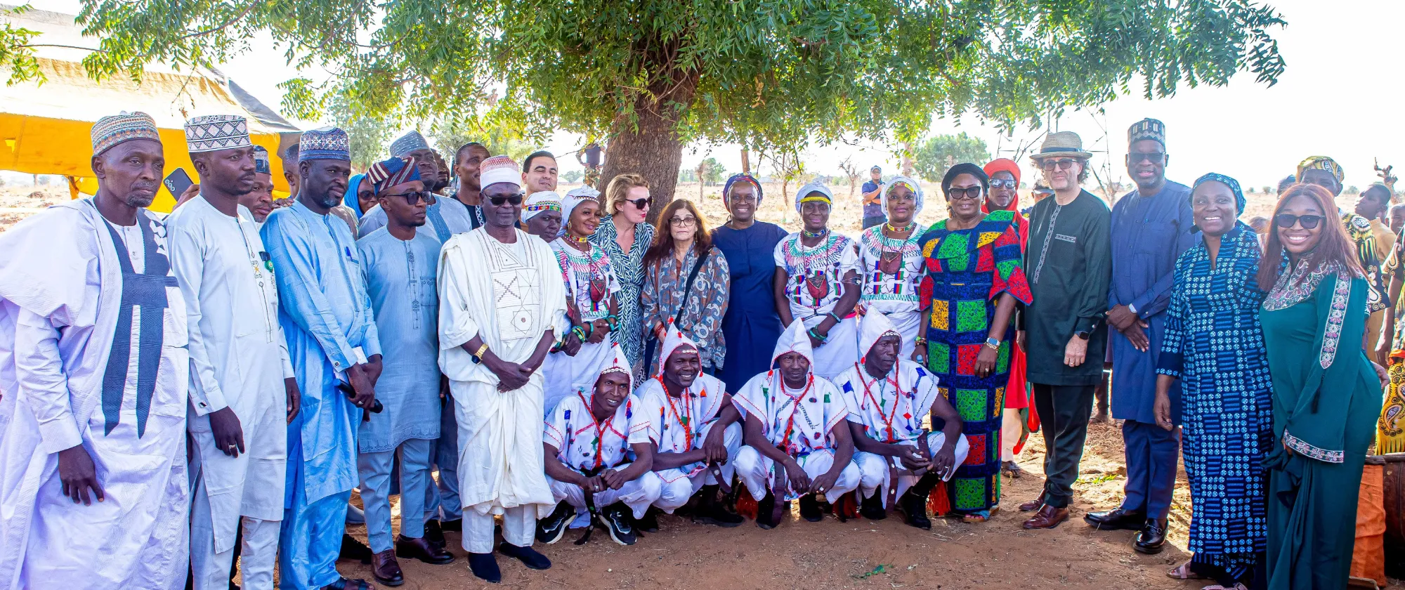 A large group of men and women in traditional and formal attire stand together outdoors under a tree for a group photograph.