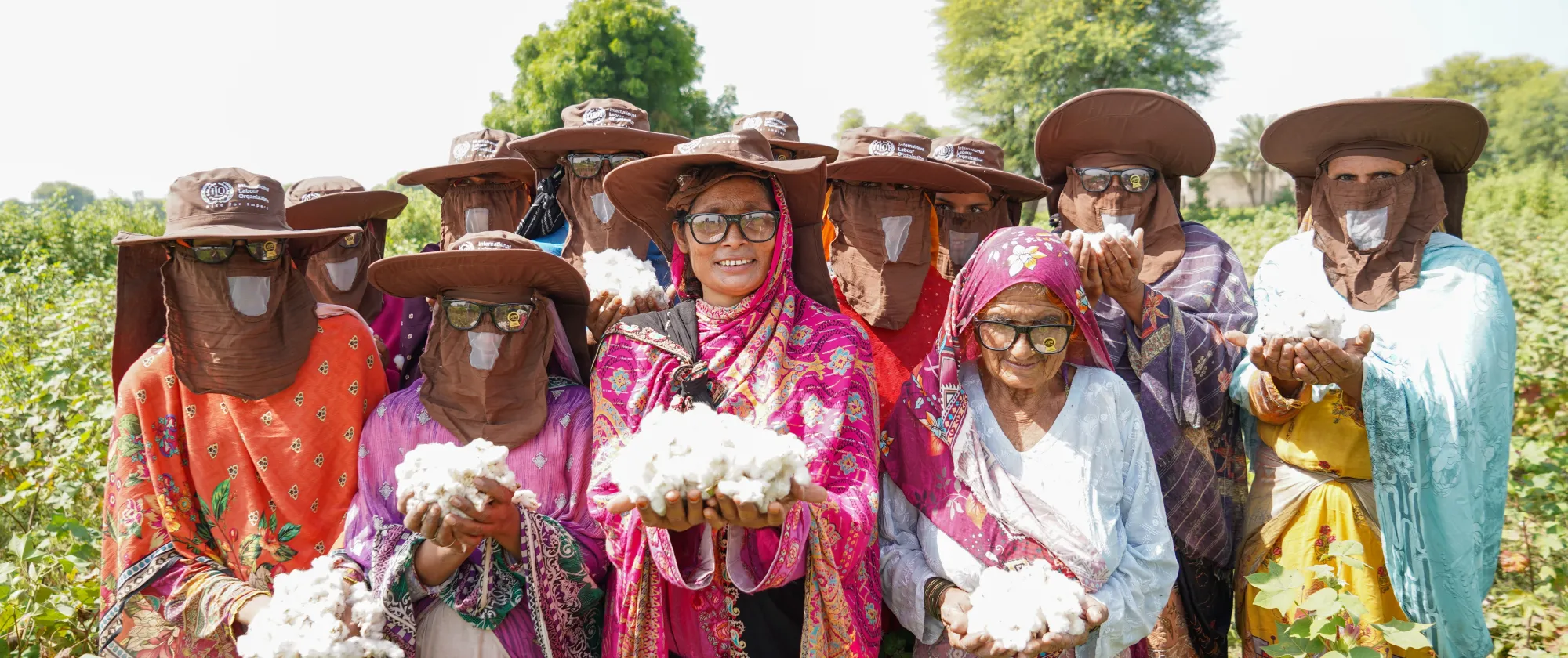 Hafeeza Mai wears a pink patterned shawl and glasses and stands in a cotton field alongside a group of women workers. They all hold tufts of cotton and wear protective broad-brimmed hats with the ILO logo on it. (Pakistan 2025)