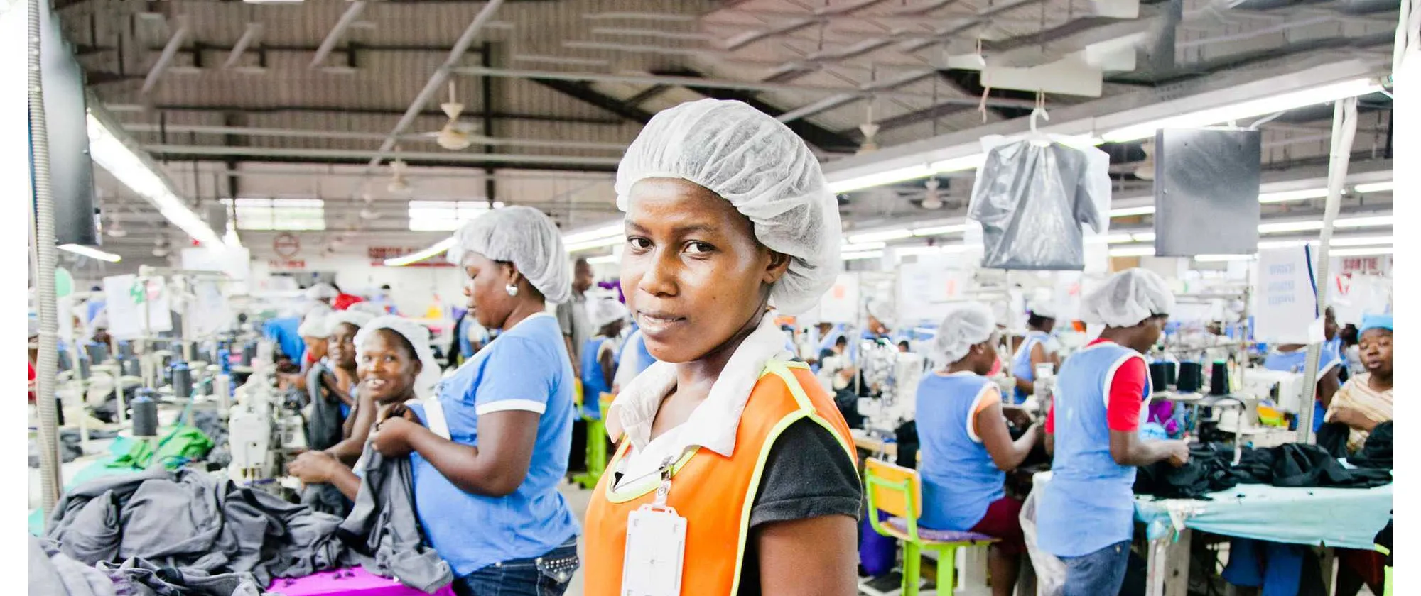 A worker at a garment factory in Haiti looks to camera, representing the ILO's Better Work Haiti programme, which supports improved working conditions in the country's textile and apparel sector. Fellow workers are visible at sewing stations in the background.