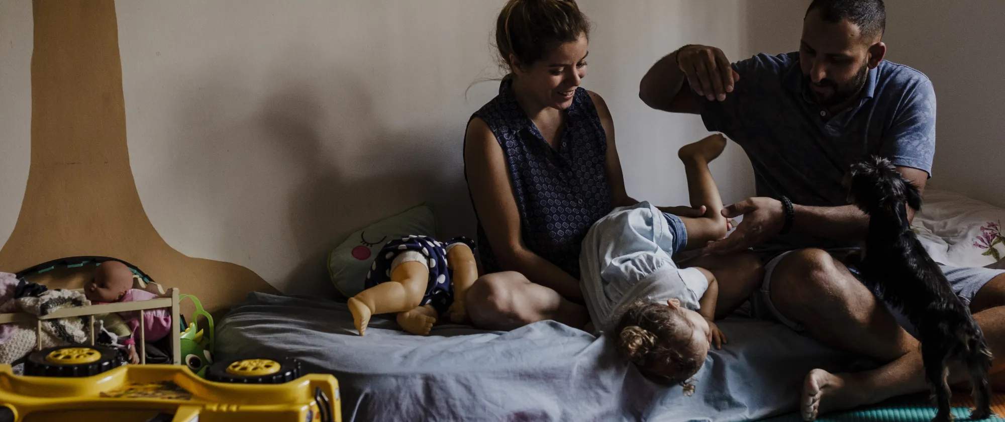 Una familia juega con su hija pequeña en una habitación infantil. La madre y el padre están sentados en el suelo sobre una colchoneta de colores mientras levantan y hacen reír a la niña. A su alrededor hay juguetes, incluyendo un camión amarillo volcado, muñecas y un perrito pequeño que se acerca curioso.