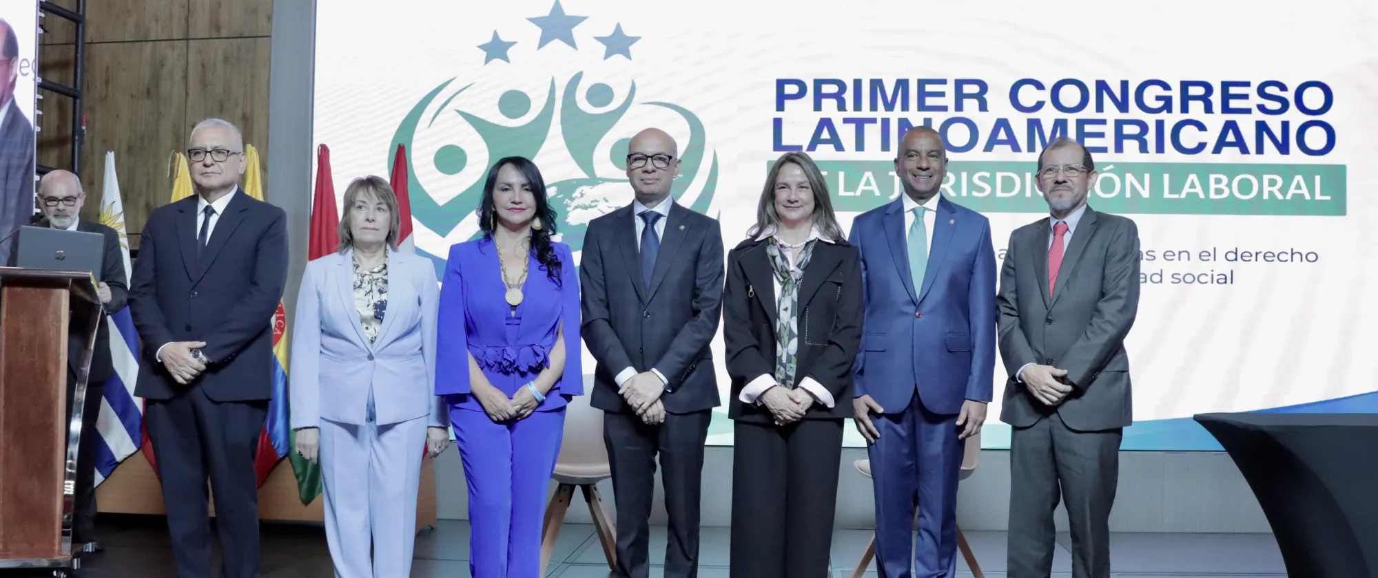 Siete personas vestidas formalmente posan en un escenario; detrás, una pantalla grande muestra el texto “Primer Congreso Latinoamericano: La Jurisdicción Laboral” junto a logotipos institucionales y banderas.