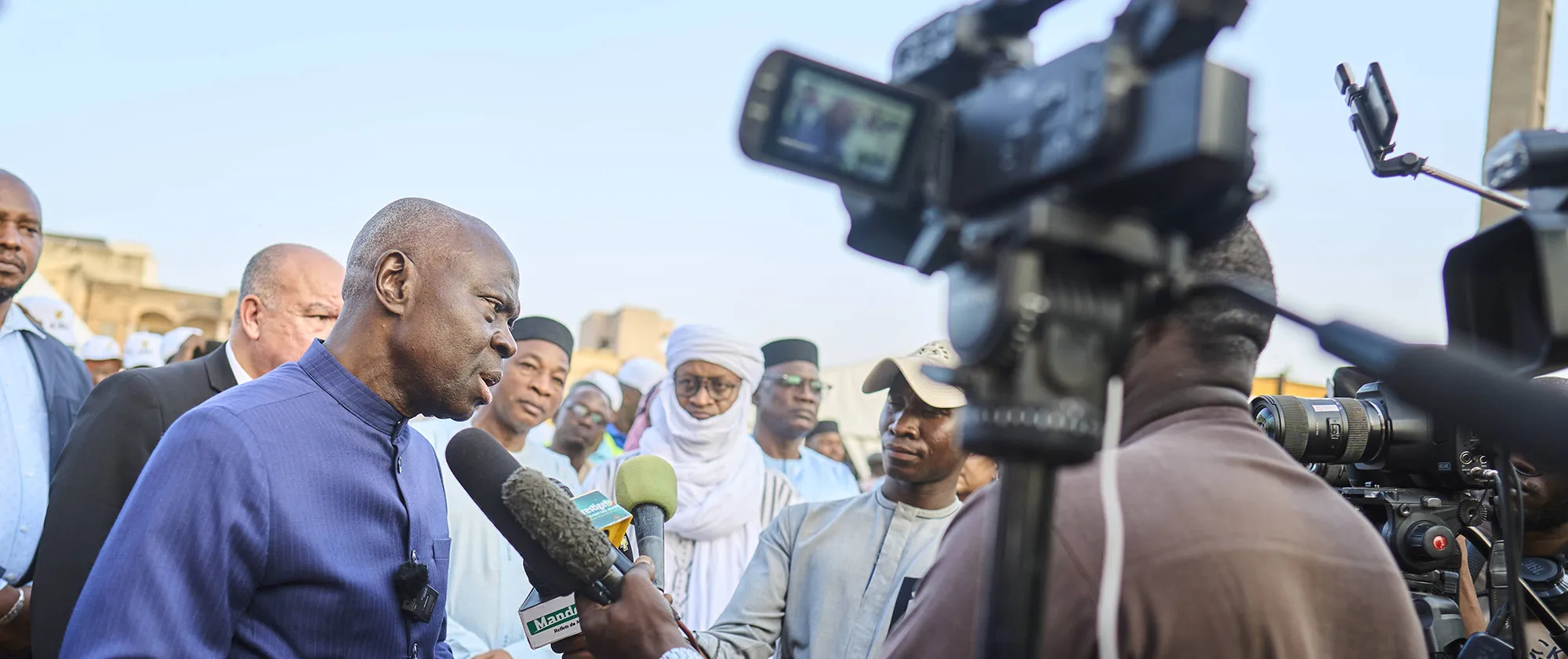 Le Directeur général de l’OIT, Gilbert F. Houngbo, s’exprime devant des journalistes lors d’une interview, avec des microphones et des caméras visibles, tandis que des responsables et des personnes présentes se tiennent derrière lui.