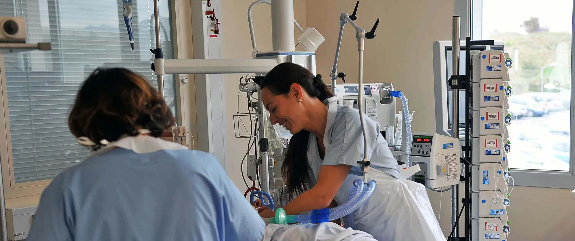 Two nurses in hospital scrubs care for a patient lying in a bed in an intensive care unit, adjusting medical equipment and monitoring devices in a clinical room.