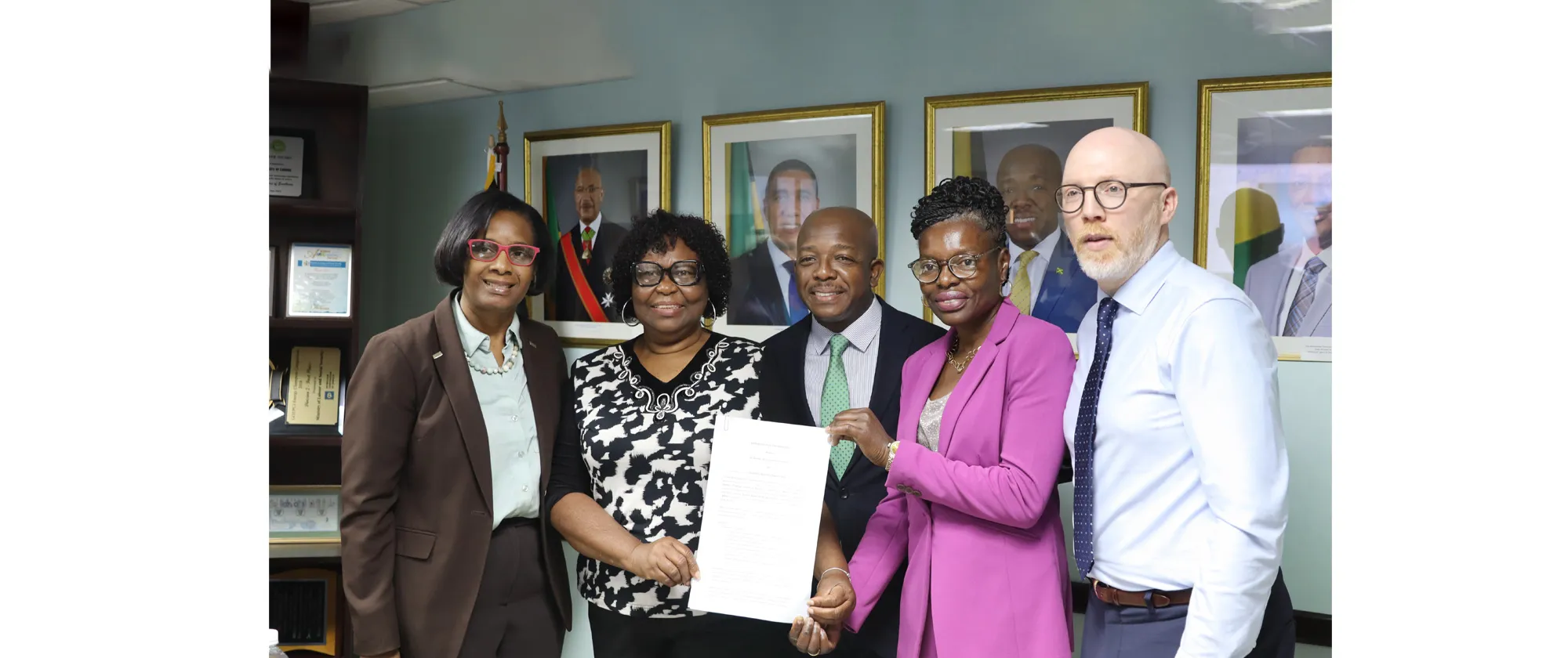 Alt Text: Signing of the MOU between ILO and JHWU  A professional group portrait features five individuals standing together in an office setting, holding a signed document. The group consists of three women and two men, all dressed in formal business attire.