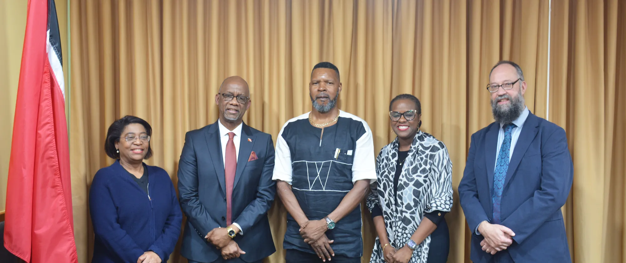 A formal group featuring five individuals standing side-by-side against a backdrop of draped gold-colored curtains. The group includes three men and two women, all dressed in professional or formal attire. The national flag of Trinidad and Tobago (red with a black diagonal stripe bordered by white) is visible on the far left.