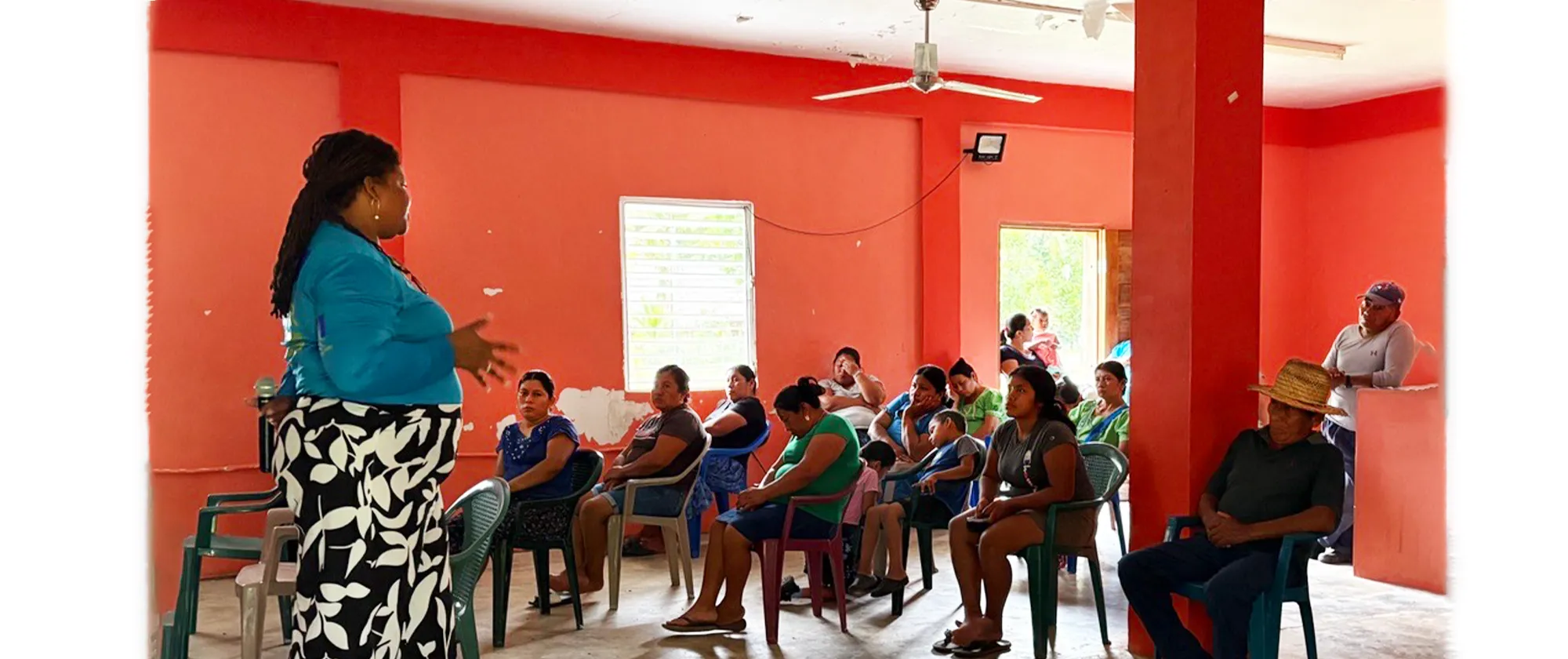 A woman stands in the foreground on the left, facing an audience and gesturing with her hands as she speaks during a community consultation. She has long dark locs and is wearing a bright blue long-sleeved top and a black-and-white floral patterned skirt.
