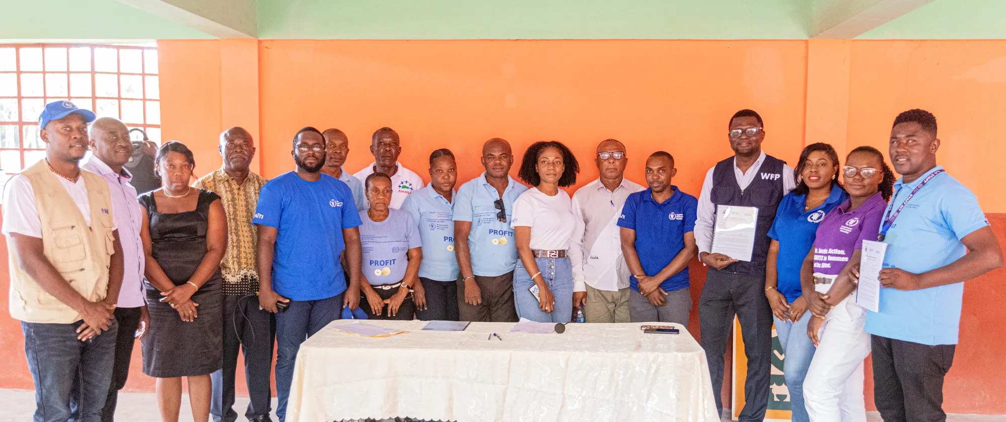 A wide group photograph shows approximately 16 people standing behind a table covered with a white tablecloth in a semi-outdoor setting. The individuals, primarily of African descent, are a mix of men and women of various ages.