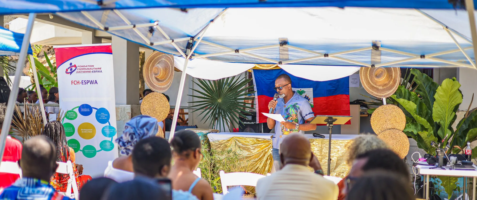 A man with a grey beard and sunglasses, wearing a blue short-sleeved shirt with a tropical print, stands behind a gold-draped table. He is holding a microphone and reading from a document.