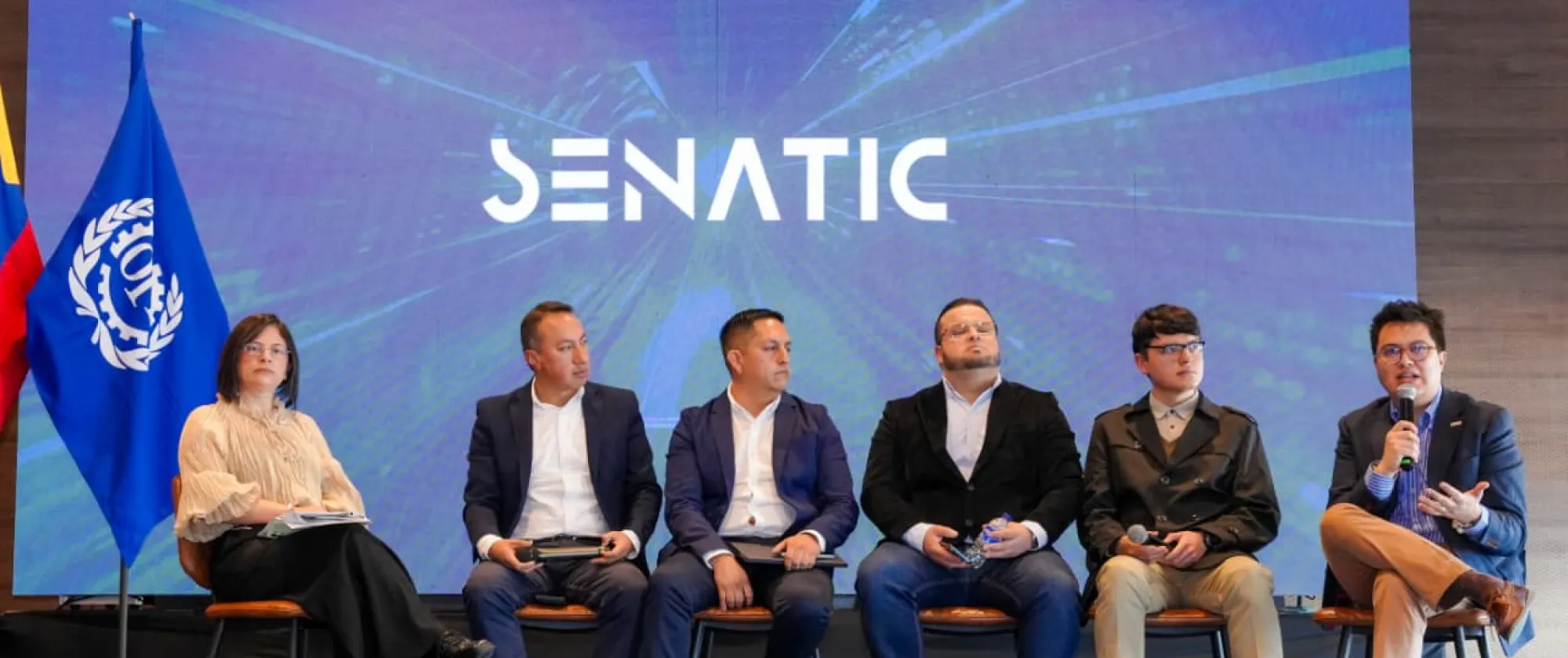Fotografía grupal en un auditorio durante evento institucional de SENATIC. Doce personas, entre representantes institucionales, invitados y participantes, posan de pie frente a una gran pantalla donde se proyecta el logo de SENATIC. Detrás de ellos se observan las banderas de Colombia y de la Organización Internacional del Trabajo, lo que denota el carácter oficial del encuentro. El ambiente transmite articulación, trabajo conjunto y compromiso con la formación tecnológica.