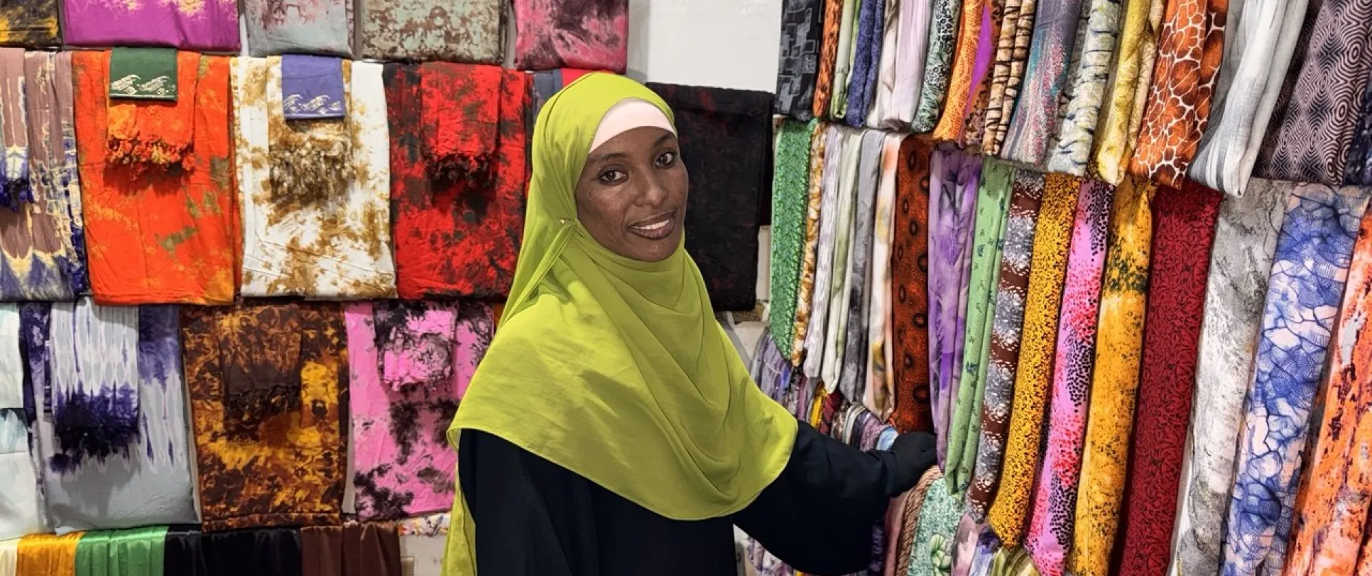 Semira Maye, an entrepreneur in Ethiopia, stands inside her textile shop surrounded by rows of brightly coloured patterned fabrics, wearing a light green headscarf and smiling while selecting material from a display.