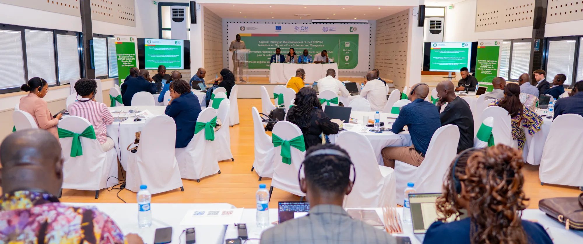 Participants seated around conference tables listen to presentations and discussions during a regional workshop on migration data management and governance in a meeting hall.