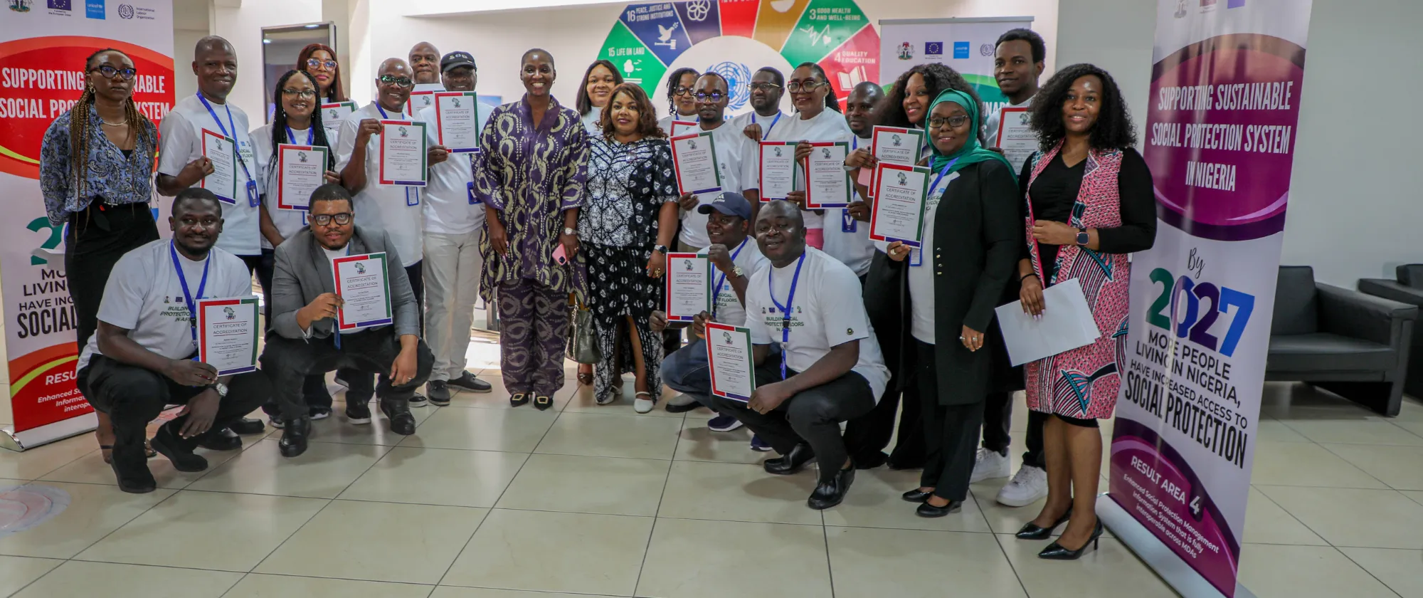 A large group of men and women holding certificates and standing together in a conference venue beside banners promoting sustainable social protection in Nigeria.