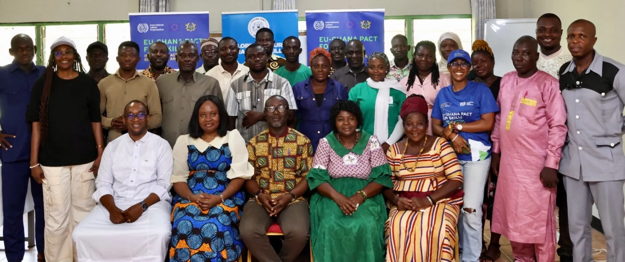 A large group of men and women standing and seated together indoors for a group photo, with ILO and EU-Ghana Pact for Skills banners displayed in the background
