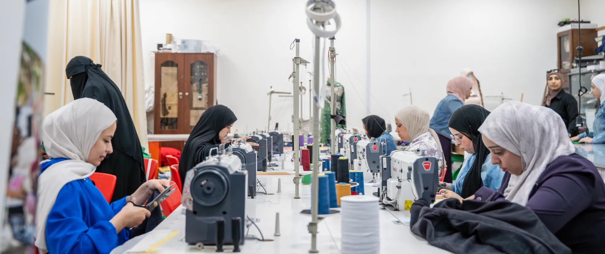 A group of women in a bright, professional vocational workshop operate industrial sewing machines and use tailoring tools to construct garments.