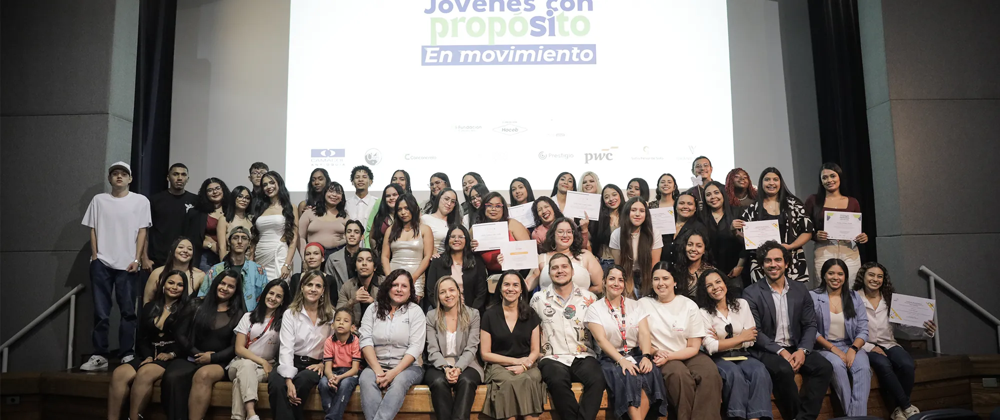 Grupo de jóvenes y representantes de organizaciones posan en un auditorio. Detrás de ellos se proyecta en una pantalla el texto “Jóvenes con propósito en movimiento” junto con varios logos institucionales. Algunas personas sostienen certificados.