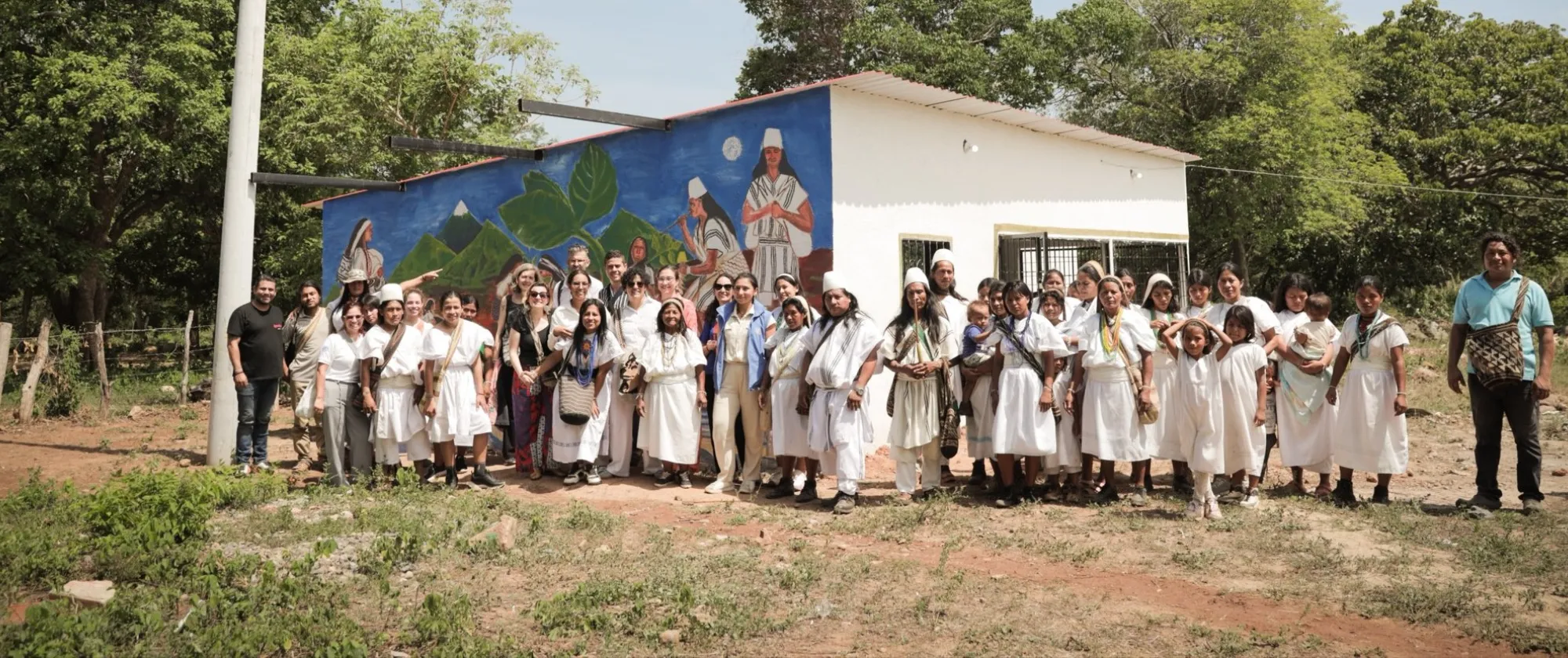 Grupo de integrantes del pueblo arhuaco y visitantes reunidos frente a una casa con un mural indígena en una comunidad rural. Muchas personas visten la indumentaria tradicional arhuaca: túnicas blancas de algodón, gorros blancos tradicionales y mochilas tejidas artesanalmente. Alrededor hay árboles y un paisaje seco bajo la luz del día.