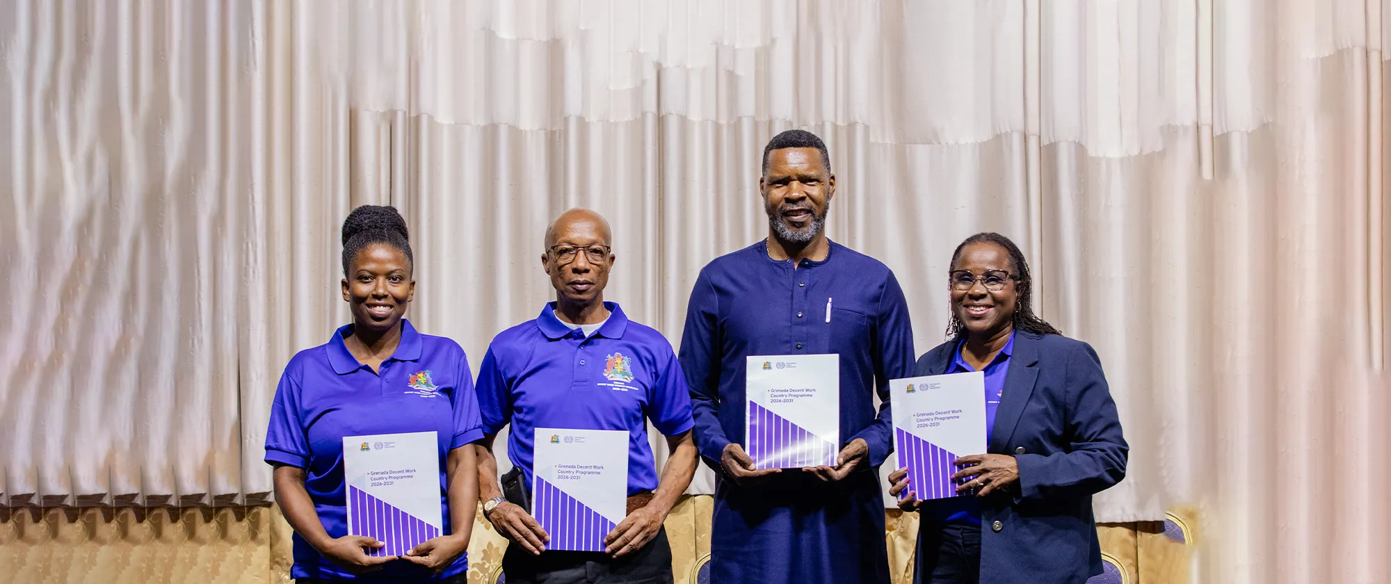 Four professionals stand side-by-side in front of beige curtains, smiling and holding up copies of the Grenada Decent Work Country Programme (DWCP) 2026–2031