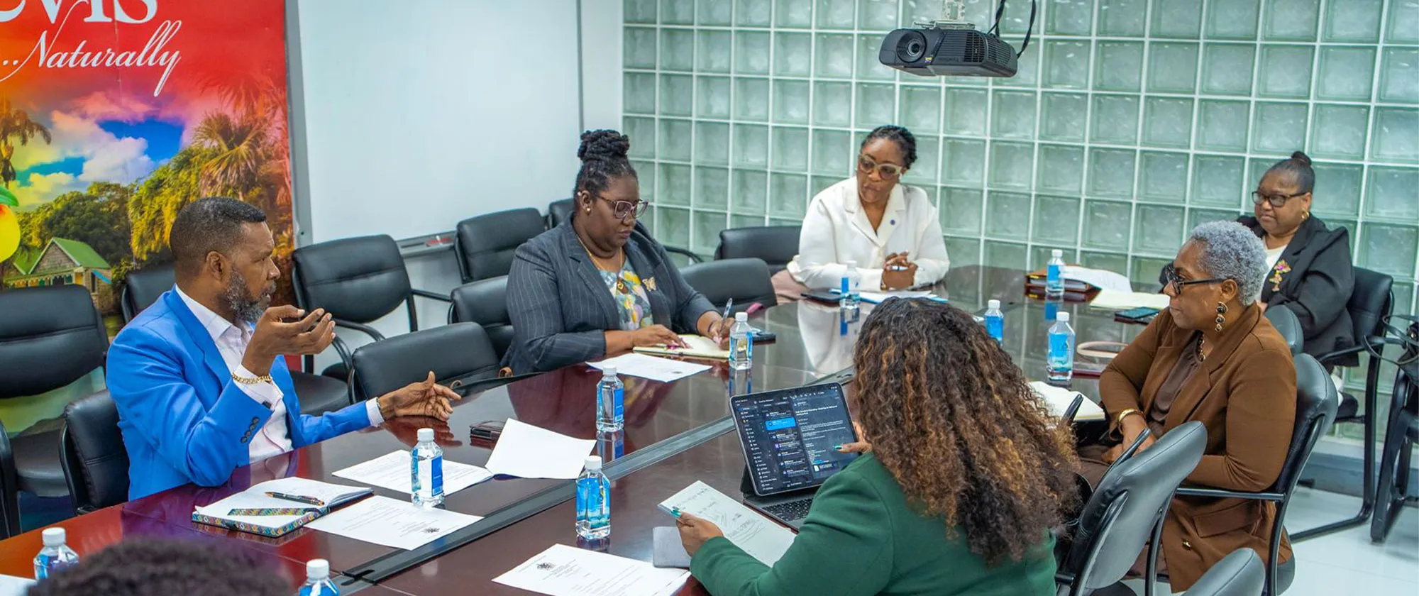 A group of professionals engaged in a meeting around a boardroom table. A man in a bright blue blazer gestures while speaking to colleagues who are taking notes.