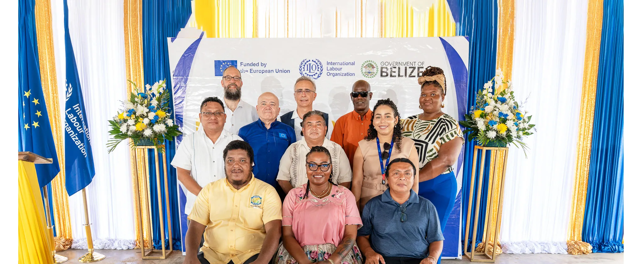 Group photo of workshop participants standing and seated in front of an International Labour Organization backdrop. The backdrop displays the logos of the European Union, the ILO, and the Government of Belize. Participants are posed indoors between blue, white, and gold draped curtains, with floral arrangements and ILO flags on either side.