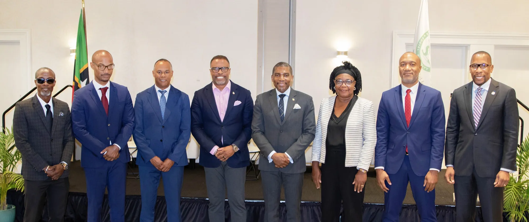 A formal group portrait of eight professionals standing in a line on a stage, flanked by the national flag of Saint Kitts and Nevis on the left and a white institutional flag featuring a green logo on the right.