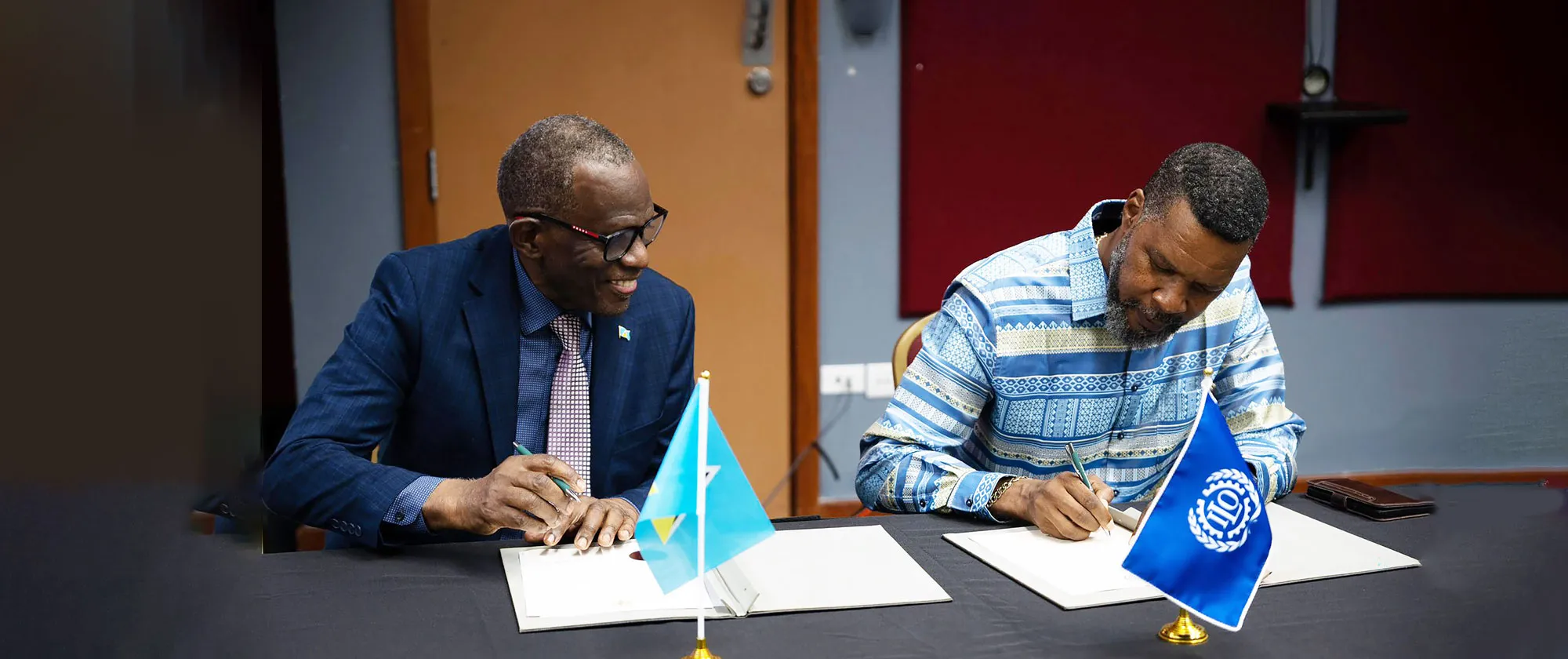 Two men seated at a table sign official documents, with the flags of Saint Lucia and the International Labour Organization (ILO) displayed in the foreground.