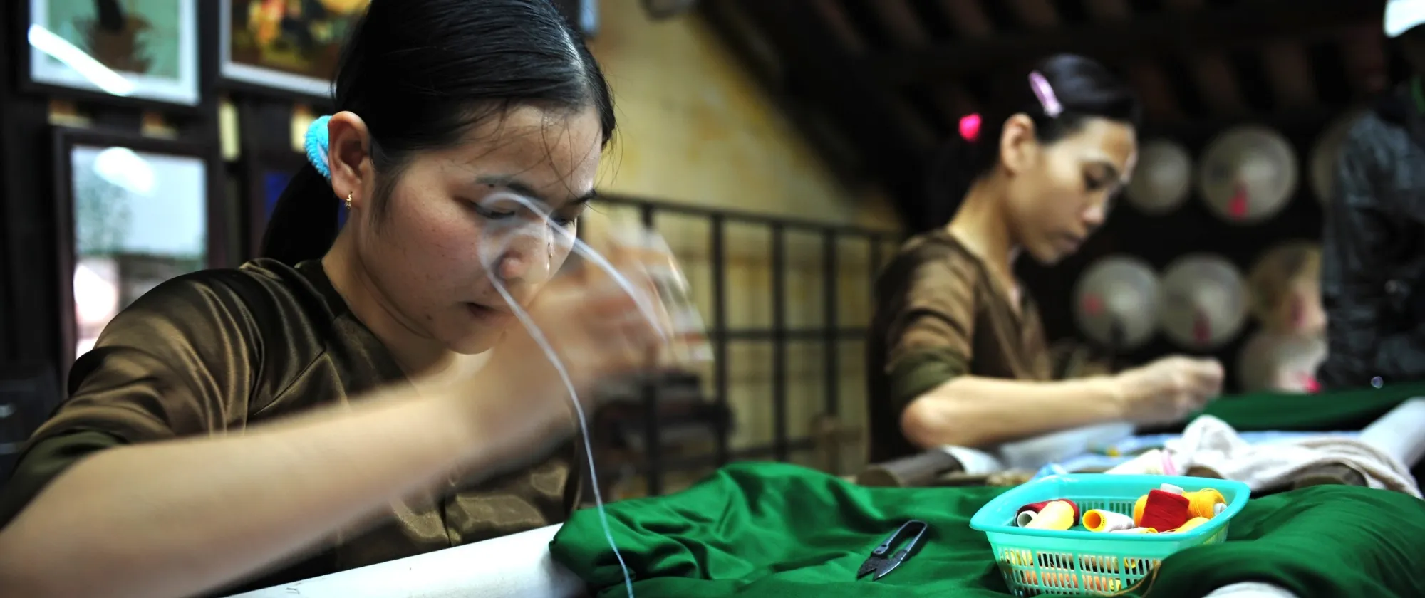 Two women sit at a worktable, carefully sewing vibrant thread into green fabric, with spools and tools arranged nearby.