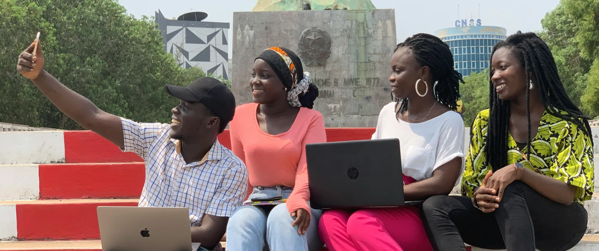 Four friends with laptops smile and take a selfie at a monument in Cotonou, Benin