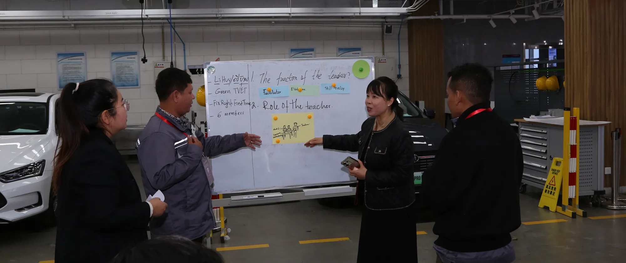 Three participants stand in front of a presentation scree, discussing content displayed on the screen in a classroom.