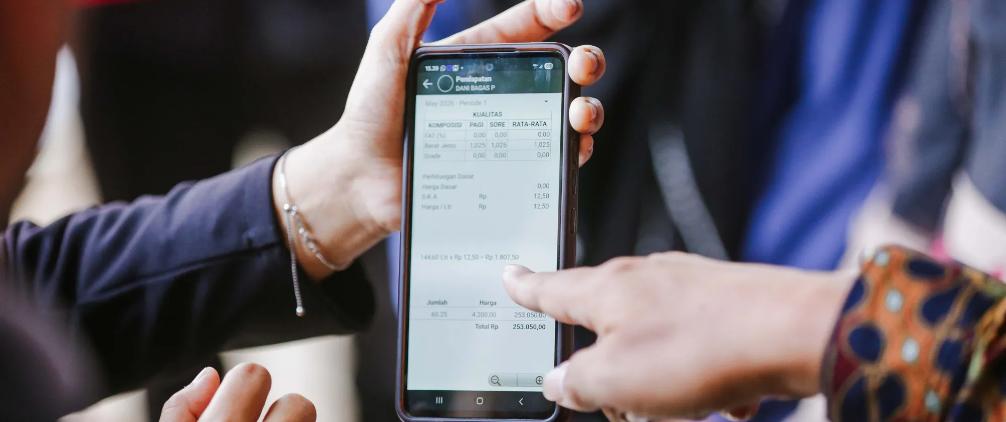 Two people review information on a smartphone displaying a dairy cooperative management application. One person points to production and payment data on the screen, illustrating the use of digital tools to improve record-keeping, transparency, and financial access for dairy farmers.