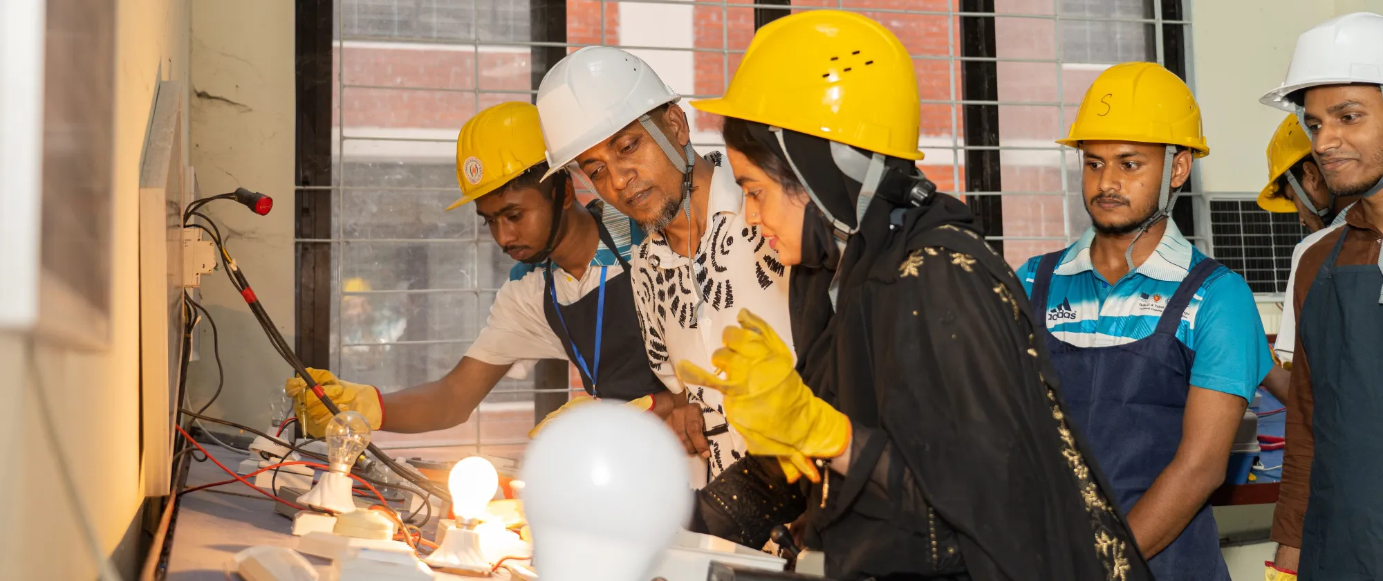 Le directeur du centre de formation technique et professionnelle, Md. Ziaur Rahman, et quatre stagiaires, dont une jeune femme au premier plan, portent des casques et des gants de sécurité et observent une installation électrique équipée de deux ampoules allumées.