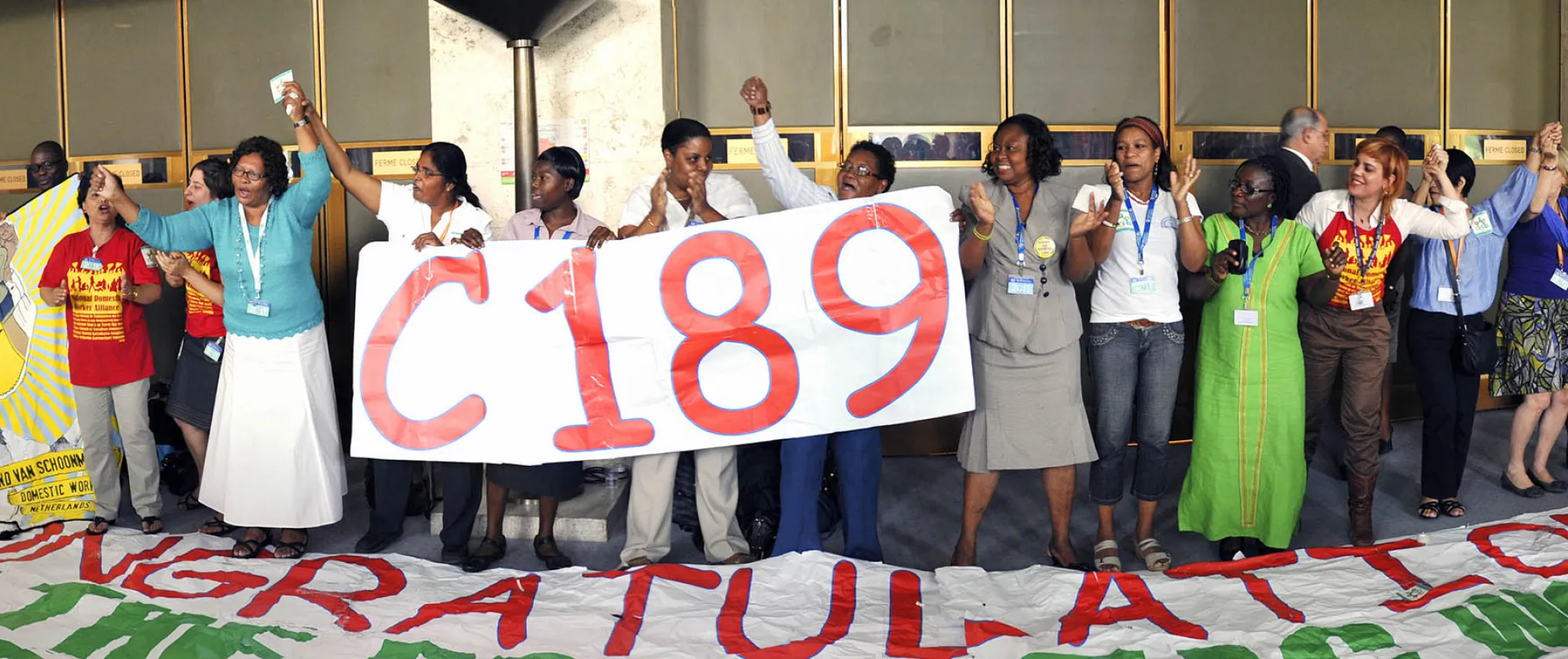 Group of domestic workers, trade union representatives and supporters standing together at the International Labour Conference, holding a large "C189" sign and a banner calling for ratification and implementation of the Domestic Workers Convention. Participants raise joined hands in celebration following the adoption of Convention No. 189 on decent work for domestic workers.