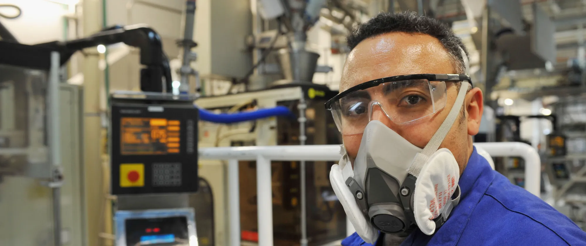 Técnico usando óculos de proteção e máscara respiratória em uma fábrica, cercado por equipamentos industriais.