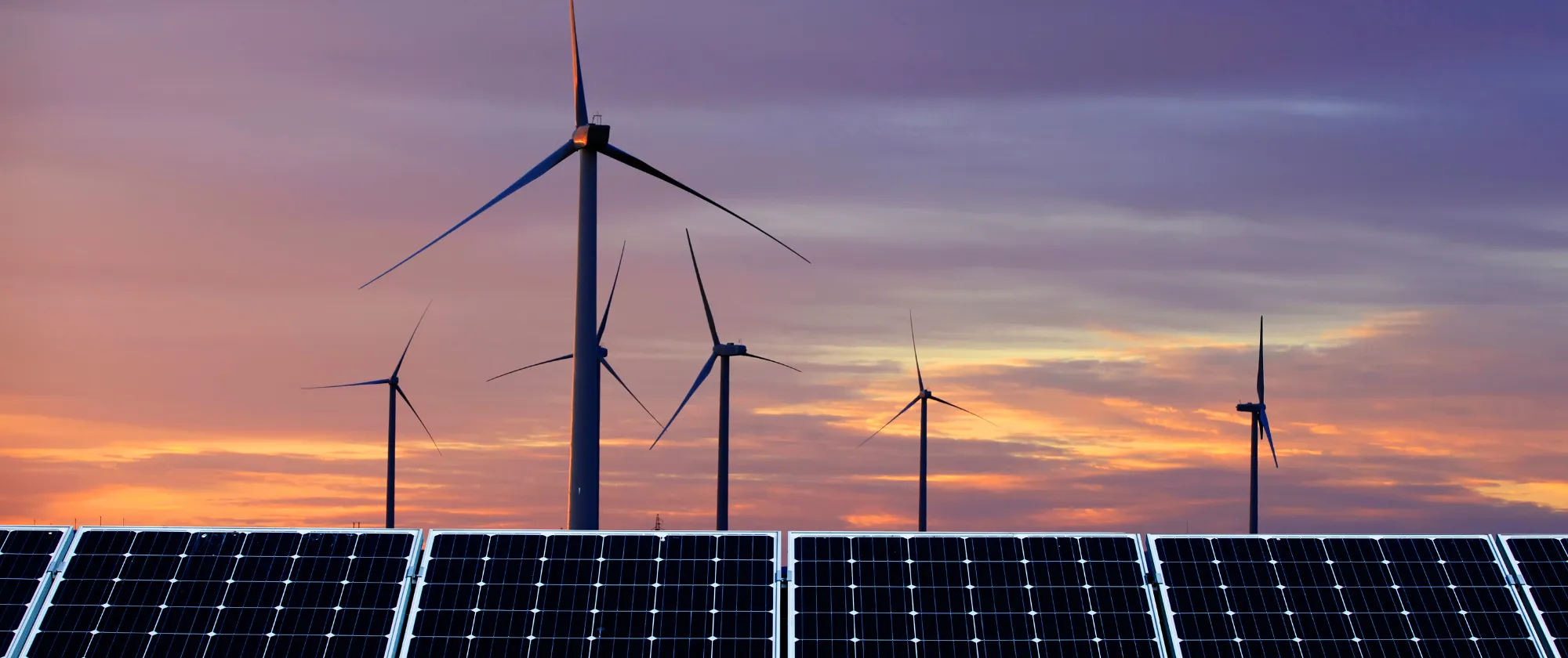 Paneles solares en primer plano y aerogeneradores al fondo durante el atardecer.