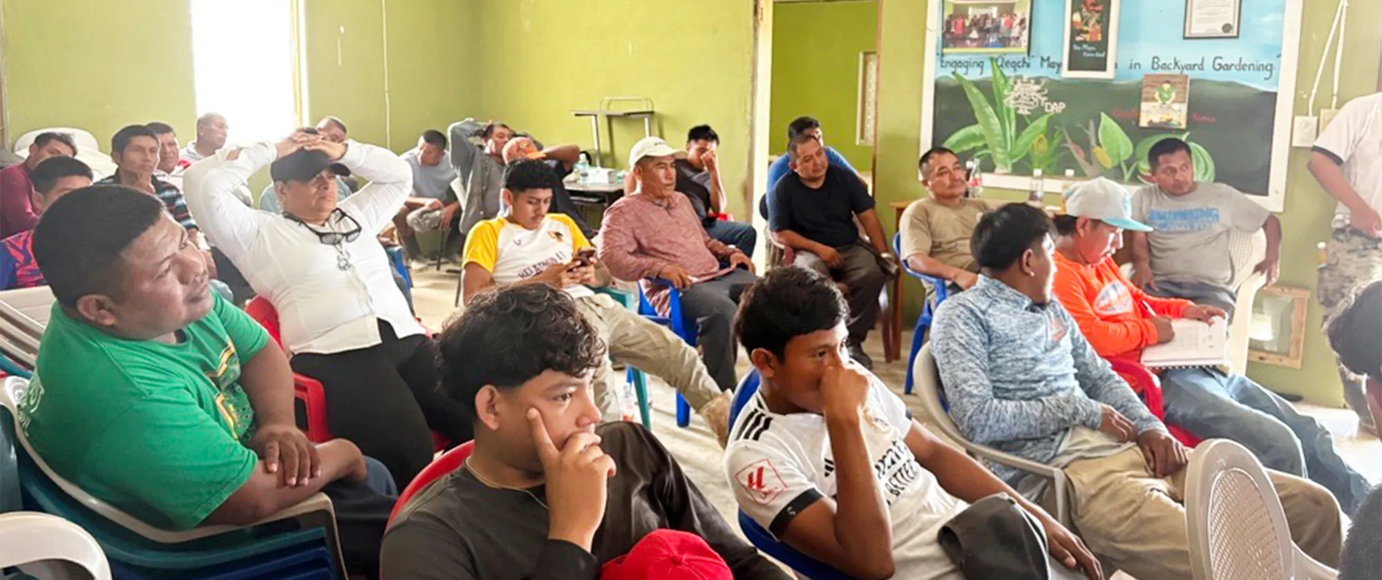 A group of adult participants seated on plastic chairs in a small community room, attentively listening during a group session.