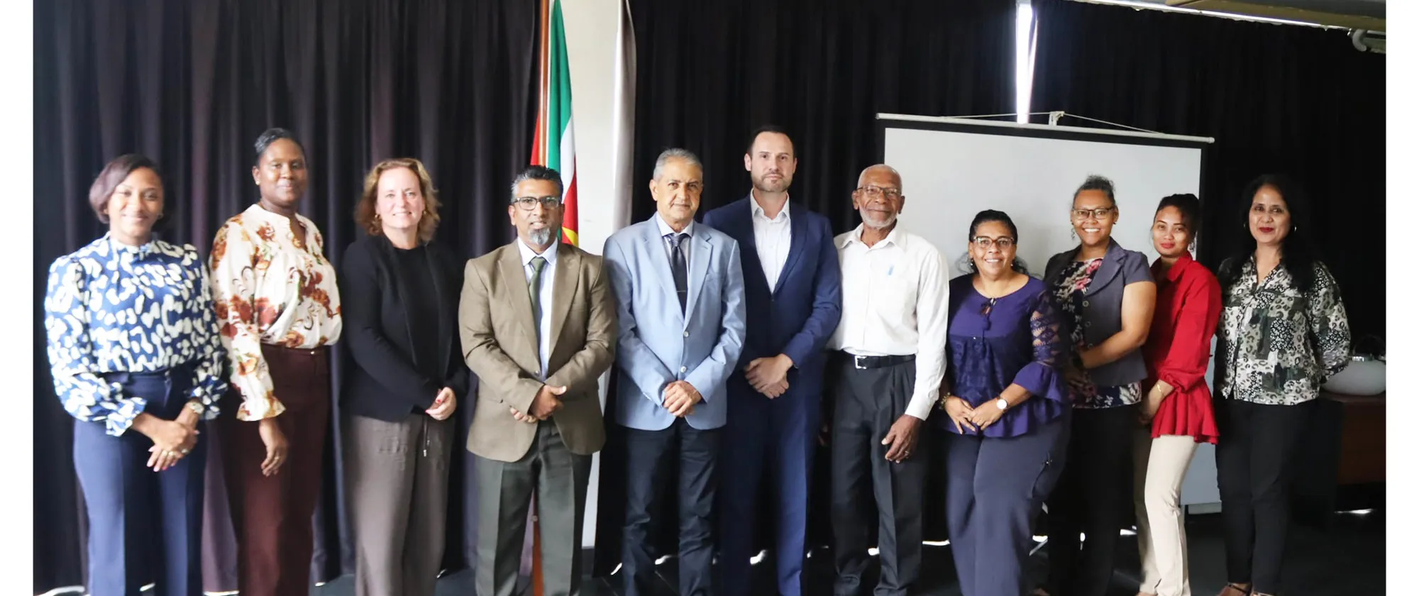 A group of eleven professionals are standing in a horizontal line for a group photo in an indoor setting. The group is positioned in front of a dark curtain backdrop, with a portion of the Suriname flag visible near the center and a blank projector screen on the right.