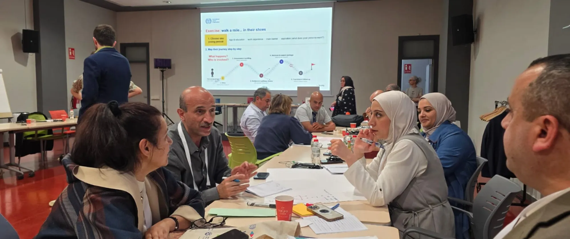 Participants seated around tables engage in small-group discussions during an interactive workshop session. Training materials, notebooks and documents are spread across the tables, while a presentation on youth pathways and service delivery is displayed on a screen in the background.