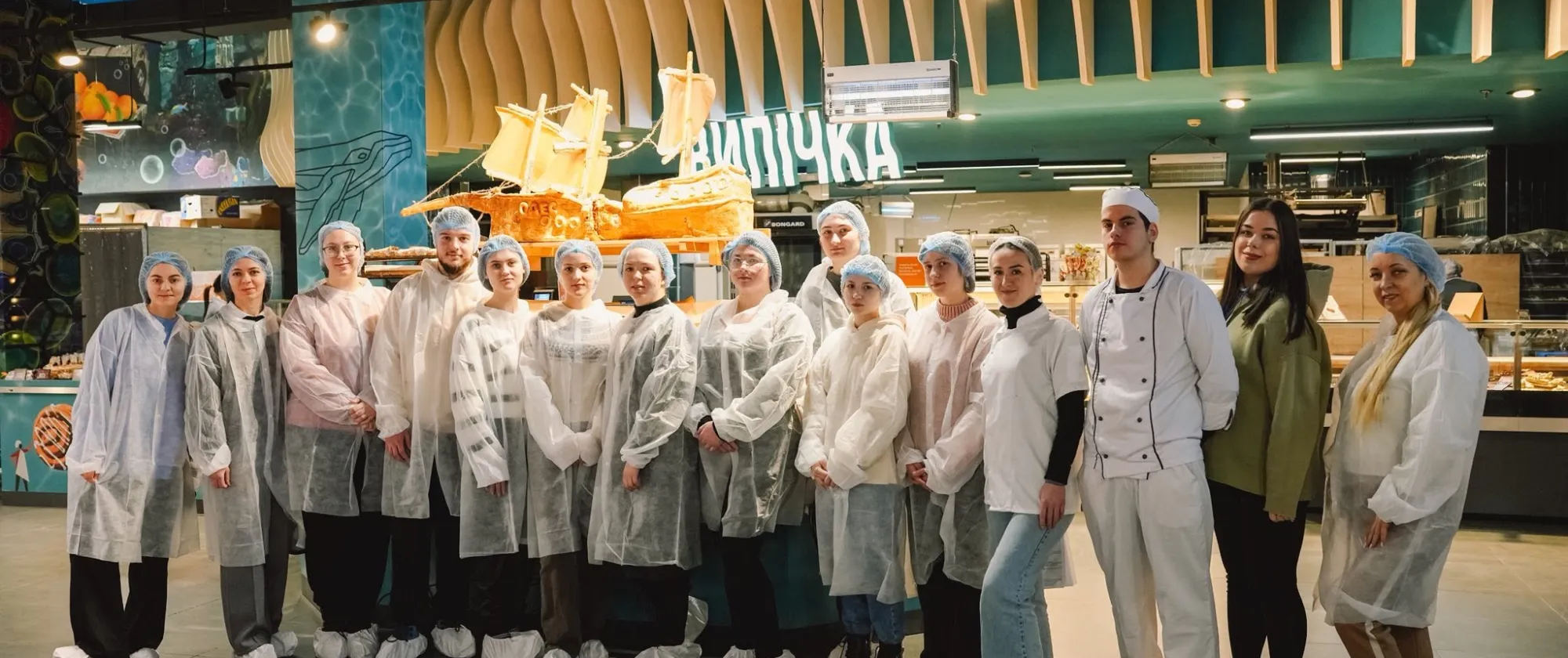 A group of students wearing protective gowns, hairnets and shoe covers stand inside a bakery production facility alongside bakery staff.