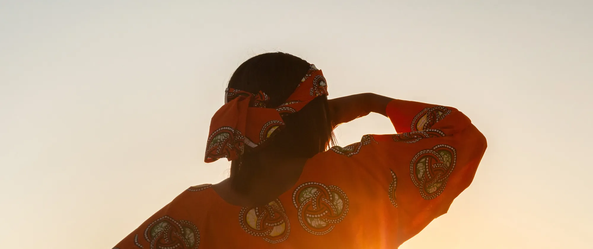 Woman in a patterned orange dress standing in a wheat field at sunset in Ghana, looking across the landscape.