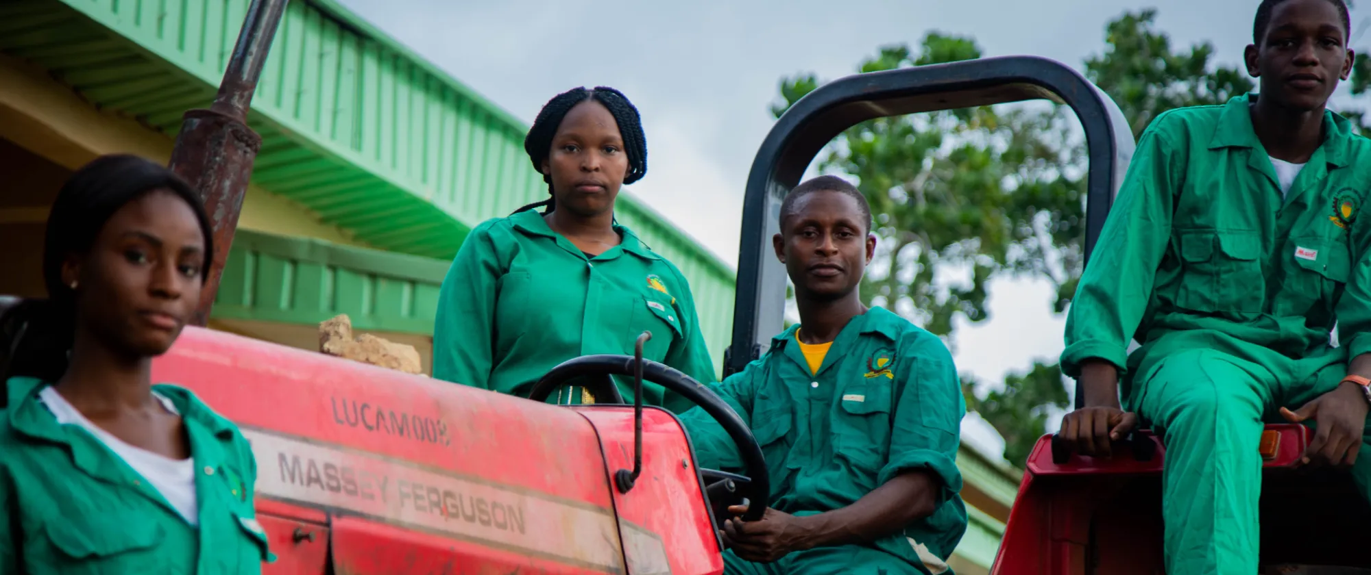 Group of young agricultural workers in green uniforms sitting on a red tractor outdoors.