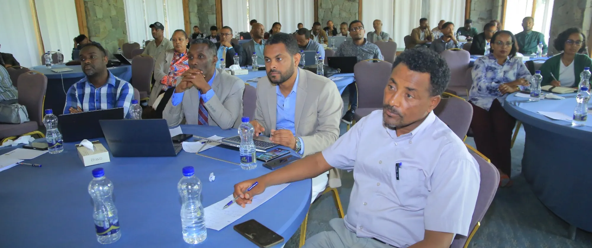 Participants seated around tables during a workshop session, listening and taking notes.